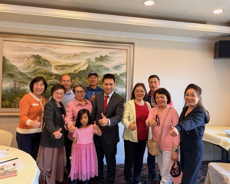 Bob Nguyen in a suit and red tie with a group of neighbors at a banquet table, everyone giving a thumbs up
