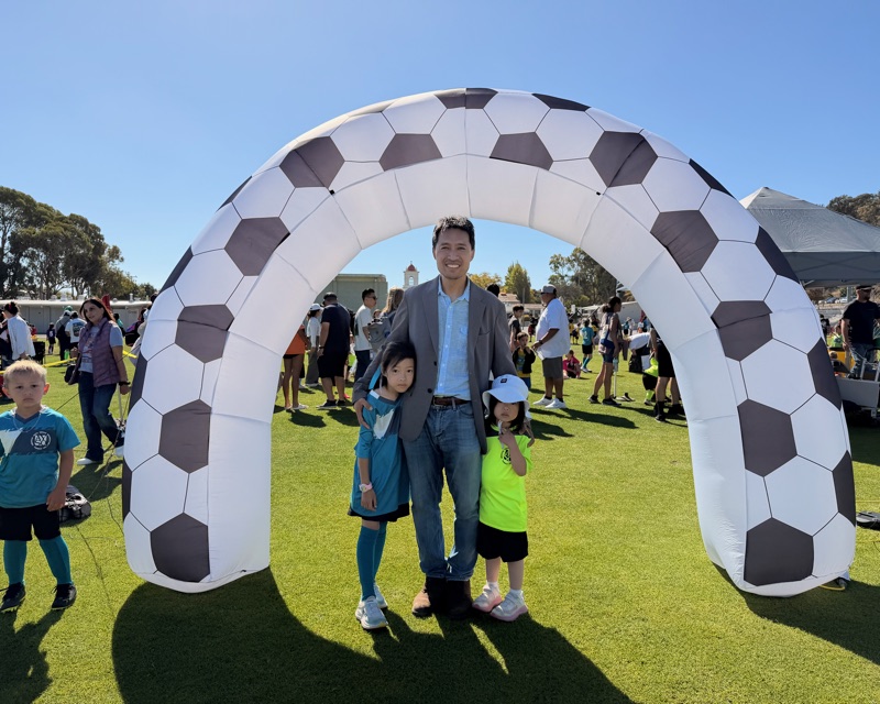 Bob Nguyen with his two daughters in their team shirts under the inflatable soccer ball arch at Millbrae AYSO opening day
