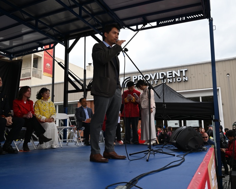 Bob Nguyen speaking into a microphone on the outdoor stage at the Millbrae Lunar New Year Festival, with seated guests behind him